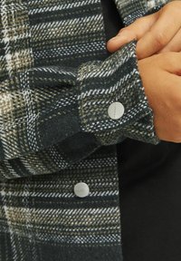 Grön och gräddfärgad rutigt flanellskjorta med knäppta manschetter, mjuk textur och silverdetaljer. Närbild på ärmdetaljer.