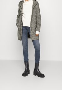 Cappotto grigio texture con cappuccio, maglione crema, jeans skinny blu e stivali neri con tacco spesso. Presenta dettagli a coste e polsini a contrasto.
