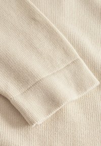 Beige stickad tyg med ribbad manschett, med en jämn textur och jämnt fördelade stygn, som visar en mjuk och lätt design.