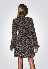 Zwarte bloemenjurk met lange mouwen, gerimpelde taille en ruches aan de manchetten. Heeft een sleutelgatdetail aan de achterkant en een losse, vloeiende vorm.