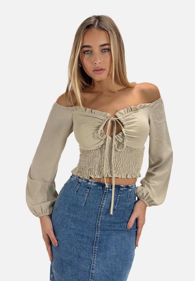 Blouse beige à épaules dénudées avec un ourlet à volants et un détail de nœud à l'avant, présentant un corps smocké et de longues manches bouffantes.