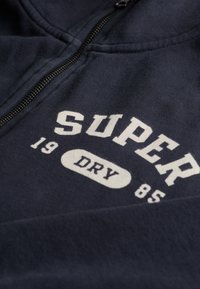 Navyblaues Zip-Hoodie aus weichem Stoff, mit weißem Drucktext: "SUPER DRY" und den Zahlen "19" und "85" darunter.