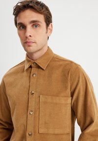 Cordura-Hemd in einem warmen Braunton, mit einem Knopfkragen, sechs Frontknöpfen und einer einzelnen Brusttasche.