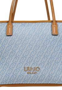 Blå stofnettaske med mønstret tekstur, håndtag i tanfarvet læder og guldfarvet præget logodetalje. Sømdekorationer på håndtagene.