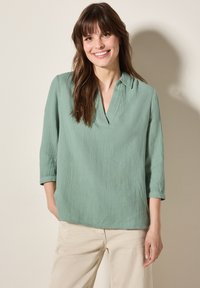 Blusa verde, con texture, a scollo a V e maniche a tre quarti. Tessuto leggero, vestibilità ampia, abbinata a pantaloni di colore chiaro.