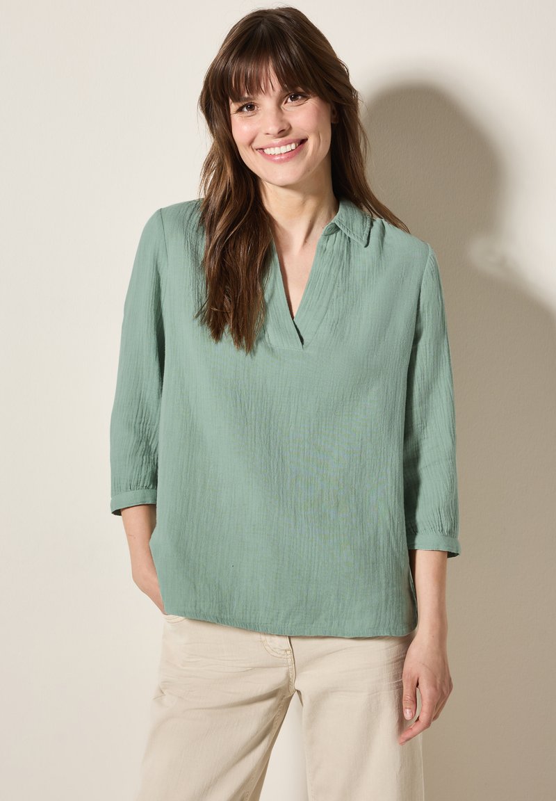 Blusa verde, con texture, a scollo a V e maniche a tre quarti. Tessuto leggero, vestibilità ampia, abbinata a pantaloni di colore chiaro.