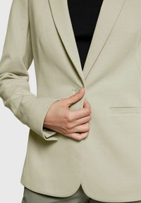 Blazer ajustado verde claro con textura suave, solapa con muesca, cierre de un solo botón y corte ajustado. Parte superior negra debajo.
