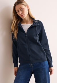 Giacca zip-up blu scuro realizzata in tessuto morbido, con colletto alto, due tasche frontali e vestibilità rilassata, abbinata a jeans in denim blu.