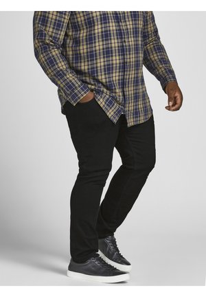 Homme portant un pantalon noir, des baskets en cuir noir avec des semelles blanches, et une chemise à carreaux verte et bleu marine à manches longues, une main dans la poche.