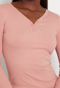 Haut à manches longues rose clair à côtes avec un col en V et des boutons décoratifs le long de l'encolure ; texture douce et coupe ajustée.