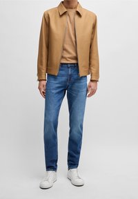 Bruine zip-up jas, tan shirt, blauwe spijkerbroek en witte sneakers. De jas heeft een kraag en zijzakken. Gladde textuur.