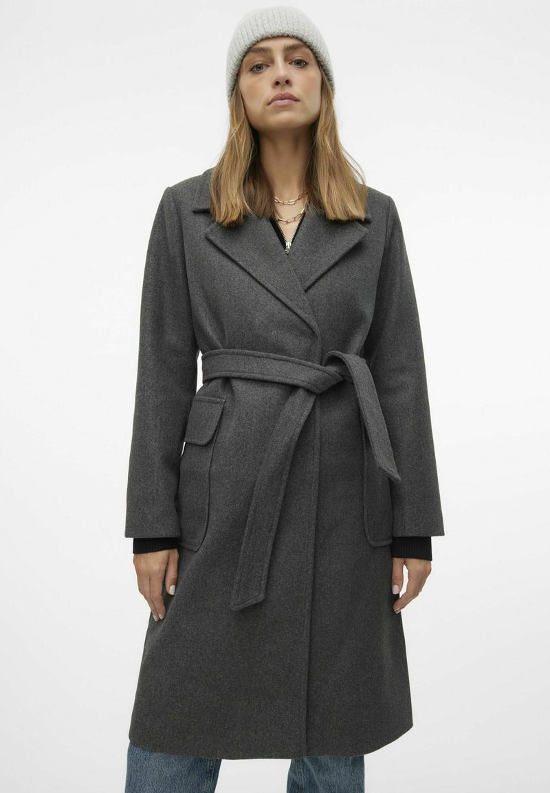 Cappotto in misto lana grigia con vita alta e cintura, rever a gola a forma di V e tasche applicate. Presenta maniche lunghe e una vestibilità dritta, abbinato a un berretto lavorato a maglia.