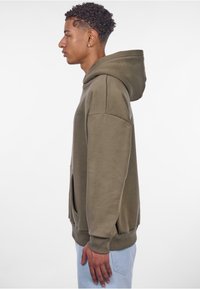 Oliven grøn oversized hoodie, blødt stof, kængurulomme, ribbede manchetter og kant, med en hætte med snor. Sideprofil.