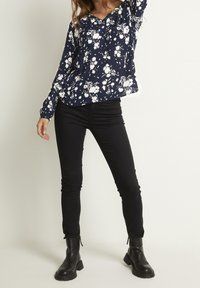 Femme portant un chemisier bleu marine à fleurs à manches longues, un pantalon skinny noir et des bottines noires, se tenant debout avec un bras levé.