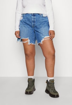 Person iført slidte blå denimshorts, hvid langærmet skjorte, hvide sokker og olivengrønne snørestøvler, der står på en grå gulv.