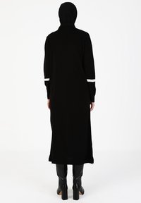 Robe noire longue avec un col montant et des manches longues, ornée de rayures blanches sur les poignets. Le tissu semble lisse et doux.