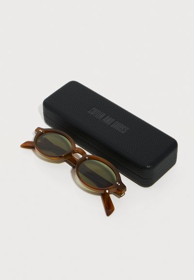 Lunettes de soleil rondes en écaille de tortue marron avec des verres foncés, à côté d'un étui rigide noir texturé avec le nom de la marque embossé.