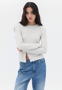 Maglia a maniche lunghe grigia chiara a coste con scollo rotondo, caratterizzata da bottoni su un lato, abbinata a jeans in denim blu.