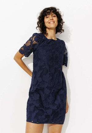 Robe en dentelle bleu marine avec des motifs floraux et des manches en maille courtes. Possède un décolleté arrondi et une silhouette ajustée.