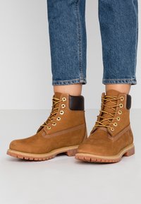 Bottes chevilles en daim marron avec une semelle en caoutchouc, un bout rond, des lacets jaunes et un col rembourré. Elles présentent une bande latérale contrastante beige et des œillets métalliques.