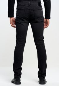 Jeans in denim nero con una vestibilità slim, dotati di tasche posteriori e una texture liscia. Abbinati a scarpe nere. Vista da dietro.