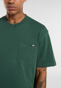 Tmavě zelené bavlněné tričko s kulatým výstřihem a krátkými rukávy, které má kapsu na levé hrudi a malou nášivku loga na kapse.