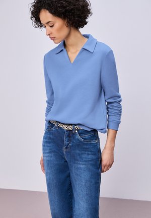 Kvinde med kort krøllet hår iført en lyseblå langærmet skjorte, blå jeans og et sort-hvidt mønsterbælte, kigger ned.