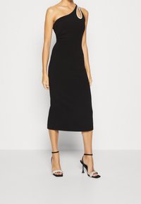 Černé midi šaty bez rukávů s asymetrickým výstřihem a kamínkovým detailem. Modelka je zkombinovala s černými sandály na podpatku se pásky.