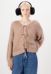 Chunky beige strikkegenser med V-hals, knytelås og lange ermer. Kombinert med blå denimjeans og svarte, fluffy ørevarmere.