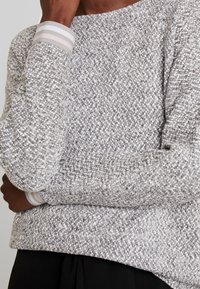 Person in einem grau-weißen strukturierten Strickpullover mit hell gestreiften Rippbündchen, die Arme über einer schwarzen Hose verschränkt.