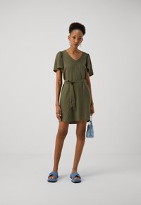 Vestido curto verde-azeitona com decote em V, mangas bufantes, cinto atado na cintura e corte reto. Combinado com sandálias azuis de deslizar e uma bolsa pequena.