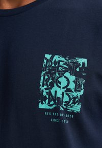 T-shirt en coton bleu marine avec un design graphique turquoise portant le texte "PETROLIND" et un motif de feuillage, accompagné des détails d'enregistrement.