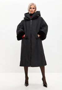 Sort oversized pufferjakke med et dobbeltlag hætte, faux fur-manchetter, lynlåslukning foran og sidelommer. Glat tekstur og knælangt design.