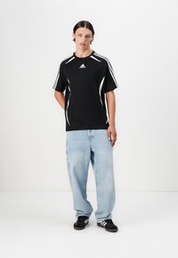 T-shirt de sport noir avec des accents blancs et trois bandes sur les manches, associé à un jean bleu clair ample et des baskets noires.