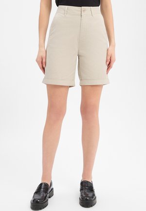 Beżowe, dopasowane szorty do kolan z podwiniętymi nogawkami, noszone z czarnymi mokasynami, pokazane na osobie stojącej na białym tle.