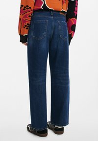 Jeans in denim blu scuro a gamba larga con una superficie liscia, dotati di due tasche posteriori e un taglio dritto, abbinati a calzature con motivo.