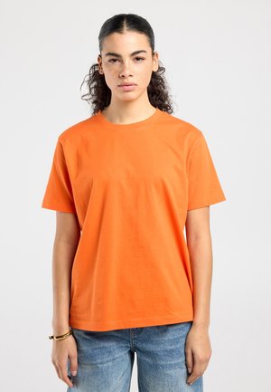 Jonge persoon met krullend donker haar, draagt een oranje T-shirt met korte mouwen en blauwe spijkerbroek, staand tegen een eenvoudige lichte achtergrond.