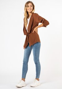 Bruine gedrapeerde cardigan met strikceintuur, gecombineerd met lichtblauwe skinny jeans en witte sneakers. Glad materiaal, driekwartmouwen.