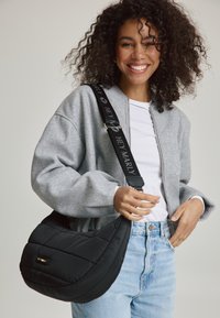 Schwarze, voluminöse Schultertasche mit geschwungener Form und gestepptem Muster, ausgestattet mit einem schwarzen Tragegurt, der mit dem weißen Schriftzug "HEY MARLY" versehen ist.