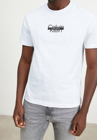 Camiseta de algodón blanca con cuello redondo y mangas cortas. Presenta un logo impreso en negro, corte ajustado y textura suave. Combinada con jeans grises.