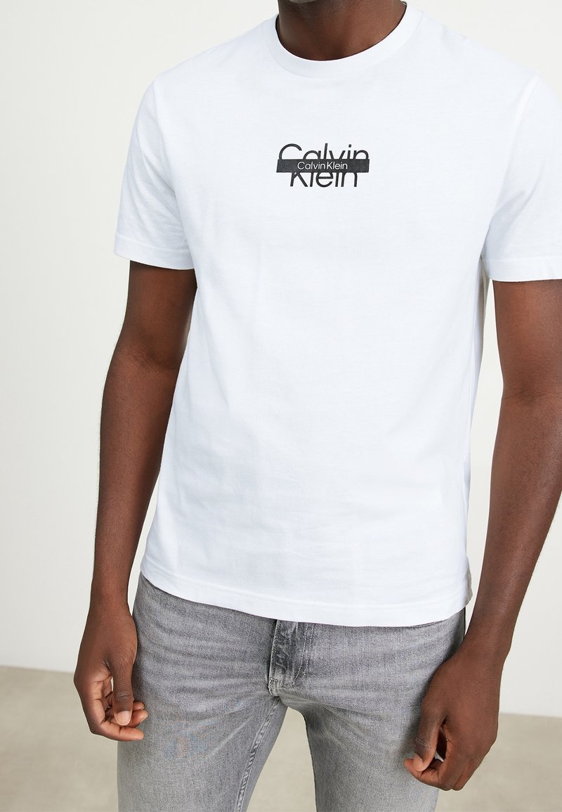 Camiseta de algodón blanca con cuello redondo y mangas cortas. Presenta un logo impreso en negro, corte ajustado y textura suave. Combinada con jeans grises.