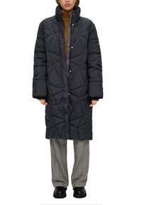 Abrigo largo negro acolchado con un alto cuello, con cierre de botones a presión. Llevado sobre un suéter marrón de cuello alto y pantalones a cuadros. Calzado negro.