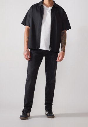 Maglietta nera a maniche corte con zip sopra una t-shirt bianca, abbinata a jeans scuri e sneaker nere, caratterizzata da un design elegante e moderno.