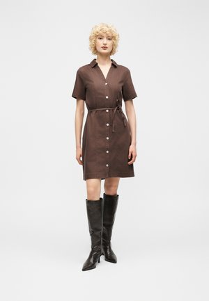 Vrouw met krullend blond haar draagt een bruin V-halsjurkje met knopen en een tailleband, gecombineerd met knielange zwarte leren laarzen.