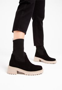 Czarne zamszowe botki za kostkę z masywną beżową podeszwą i czarną, prążkowaną górą przypominającą skarpetę, z teksturowanym wzorem na materiale.