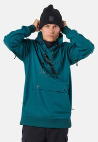 Jonge man die zijn zwarte beanie rechtzet, draagt een turquoise hoodie met een voorzak en zwarte broek, tegen een effen witte achtergrond.