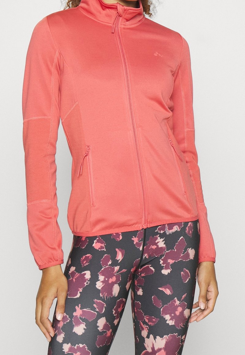 Veste zippée corail avec des panneaux latéraux texturés et deux poches avant, portée avec un legging noir à motif floral dans des teintes de bordeaux et de rose.