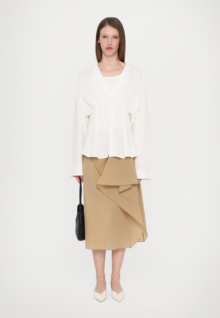 Chemisier blanc boutonné avec des manches longues, associé à une jupe enveloppante beige avec un tissu drapé. Le modèle tient un sac noir texturé.