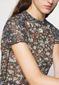 Průsvitný top s krátkým rukávem zdobený vícebarevným květinovým vzorem na tmavém pozadí, s kulatým logem Nike na výstřihu.