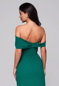 Grön off-shoulder klänning i mjukt tyg, figursydd silhuett och synliga sömdetaljer. Har en knappstängning bak.